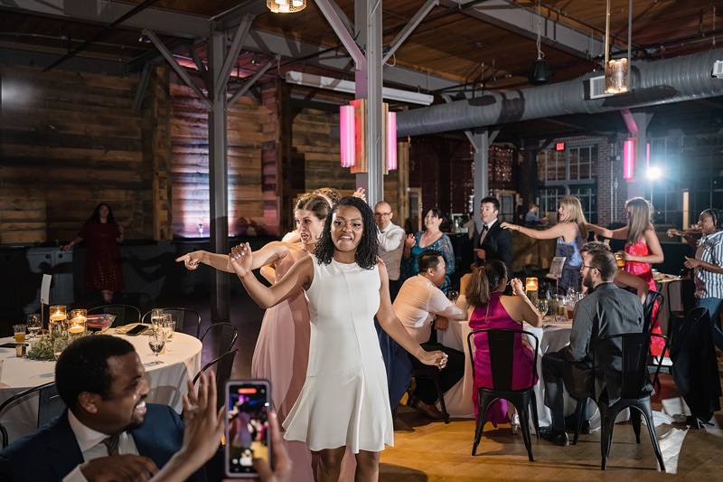 Two joyful women dance and pose for a photo at The Rickhouse wedding reception, with guests dining and dancing under warm, ambient lighting in an elegant, rustic venue.