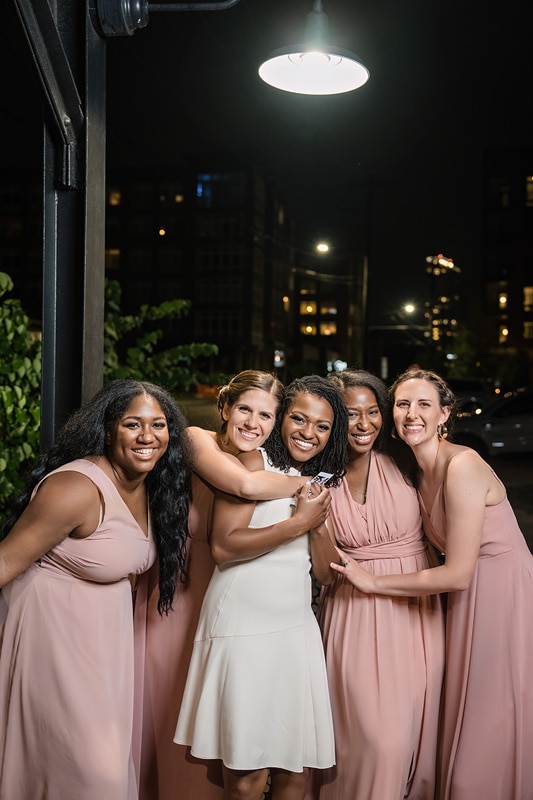 A radiant bride in white embraces her joyful bridesmaids in matching blush dresses under a warm streetlight on a blissful evening at The Rickhouse, friendship aglow amidst urban surroundings.