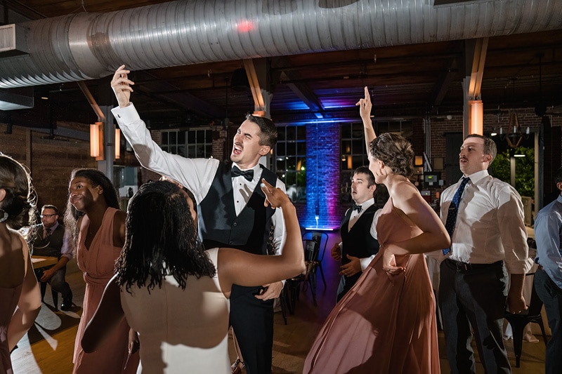 Energetic partygoers in formal attire dance and sing along at a lively The Rickhouse wedding reception, with one man enthusiastically taking a selfie amidst the joyful crowd.