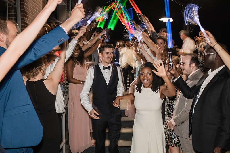 A joyful bride and groom walk through a tunnel of friends holding glowing sticks at The Rickhouse, celebrating their wedding night with smiles and cheer.