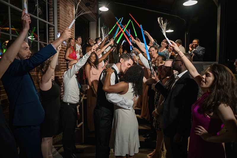 A couple shares a romantic dance under an archway of glowing sticks held by cheering guests at The Rickhouse wedding reception, surrounded by joy and warmth.