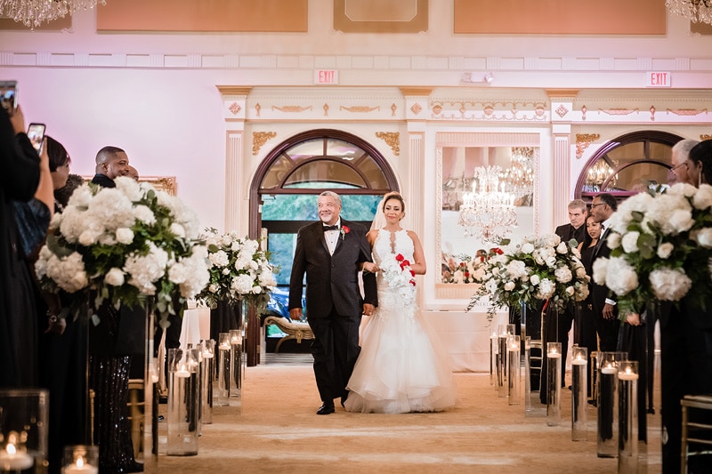 A bride walking down the aisle with an older gentleman, possibly her father, in the grand marquise ballroom as guests look on and take photos.