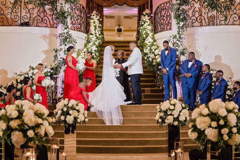 A wedding ceremony in the grand marquise ballroom with a bride and groom exchanging vows at the altar, flanked by bridesmaids in red dresses and groomsmen in blue suits.