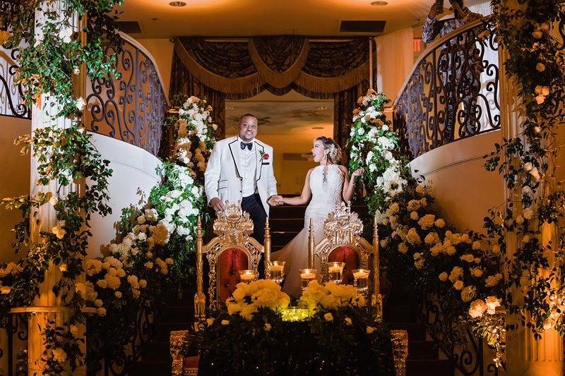 A couple holding hands at the top of a staircase adorned with white flowers at a grand marquise ballroom wedding.