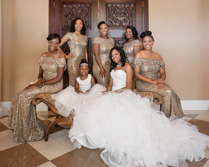 Seven smiling women in matching gold dresses, one in a white gown, seated and standing around an ornate bench at the Grand Marquise Ballroom wedding.