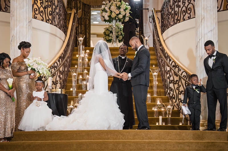 A Grand Marquise Ballroom wedding ceremony in progress with the bride and groom holding hands at the altar, surrounded by bridesmaids, groomsmen, and two young ring bearers.