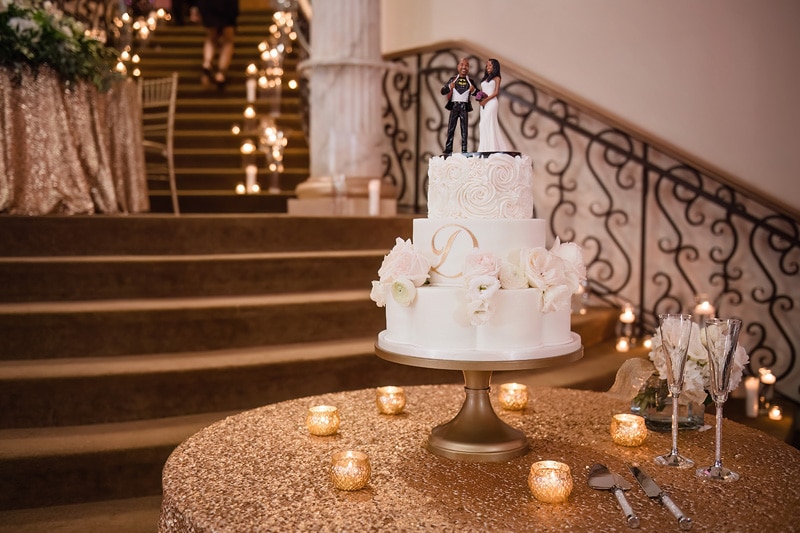 A Grand Marquise Ballroom wedding cake with figurine toppers displayed on a table with candles and floral decorations, with a staircase in the background.
