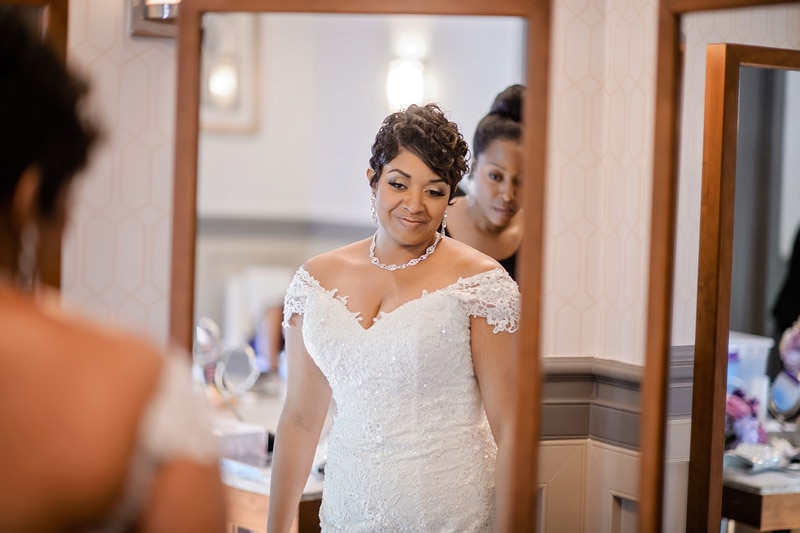 A radiant bride in a lace-trimmed gown admires her reflection at the Paramount Event Venue, accompanied by a smiling bridesmaid, in a moment of anticipation on her wedding day.