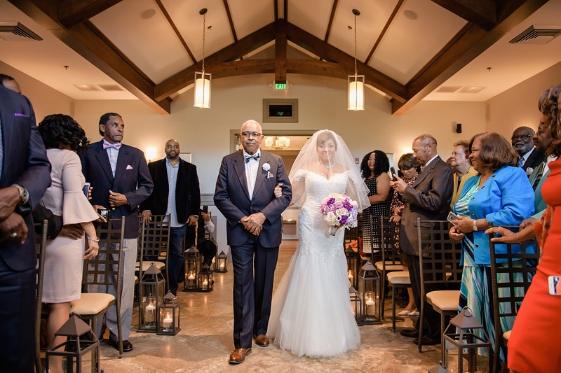 A bride walks down the aisle on the arm of an older gentleman, surrounded by guests standing in reverence at a warmly lit Paramount Event Venue wedding ceremony.