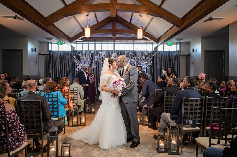 A joyous couple shares their first kiss as newlyweds while surrounded by warmly lit lanterns and guests at their elegant Paramount Event Venue wedding ceremony inside a hall with rustic wooden beams overhead.