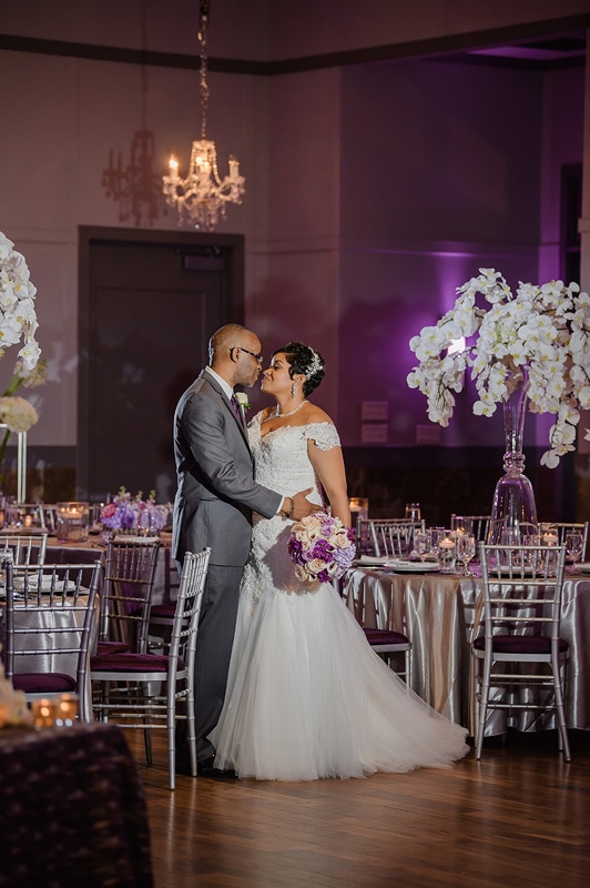 A couple shares an intimate dance in the Paramount Event Venue, a lavishly decorated ballroom with purple lighting, crystal chandeliers, and elegant floral arrangements, celebrating their wedding day surrounded by love and