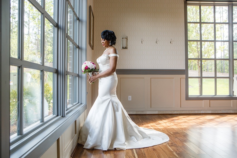 A radiant bride in an elegant white gown, holding a bouquet, gazes thoughtfully out a sunlit window at the Paramount Event Venue, savoring a quiet moment before her wedding ceremony.
