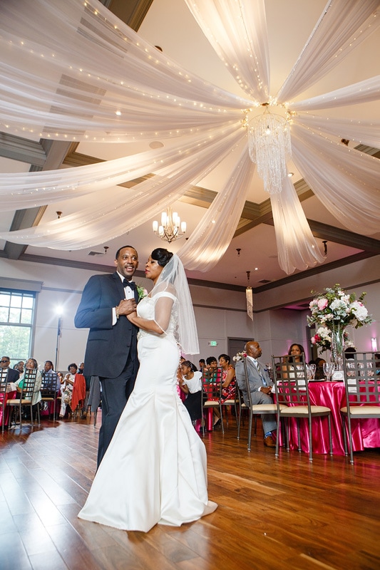 A joyful couple shares their first dance under elegant drapery and chandeliers at the Paramount Event Venue, surrounded by friends and family, capturing a moment of love and celebration at their wedding reception.