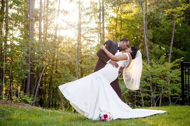 A newlywed couple shares a romantic dip kiss on a lush lawn at the Paramount Event Venue, backlit by the golden rays of a setting sun filtering through trees, highlighting the bride's flowing gown and