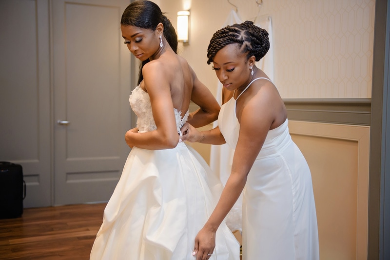 A bridesmaid carefully fastens the back of the bride's elegant white wedding dress, both women looking focused on the task at the Paramount Event Venue, set against a neutral-toned room.