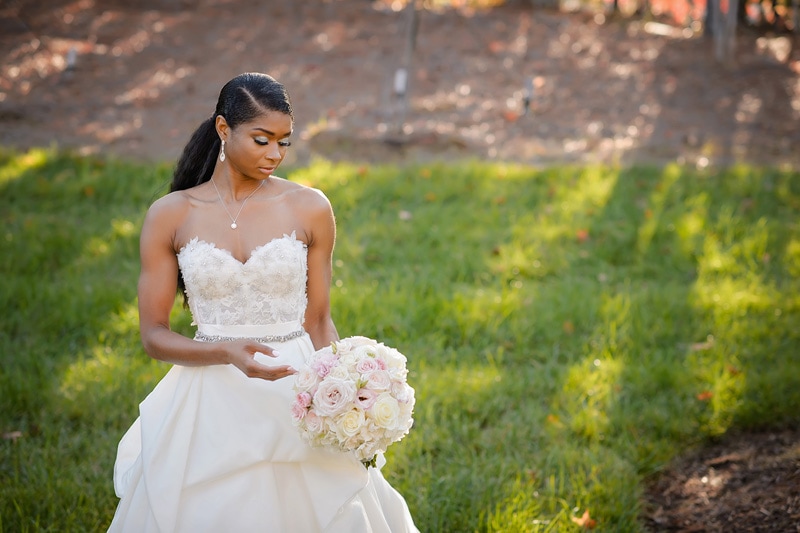 A bride in an elegant strapless gown holds a delicate bouquet of pale pink roses, lost in a moment of reflection amidst a serene outdoor setting at the Paramount Event Venue.