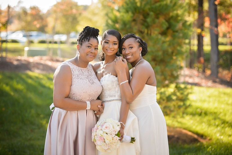 Three women dressed in formal attire, smiling at an outdoor Paramount Event Venue wedding, with one in a wedding gown holding a bouquet, suggesting a celebration of marriage on a sunny day.