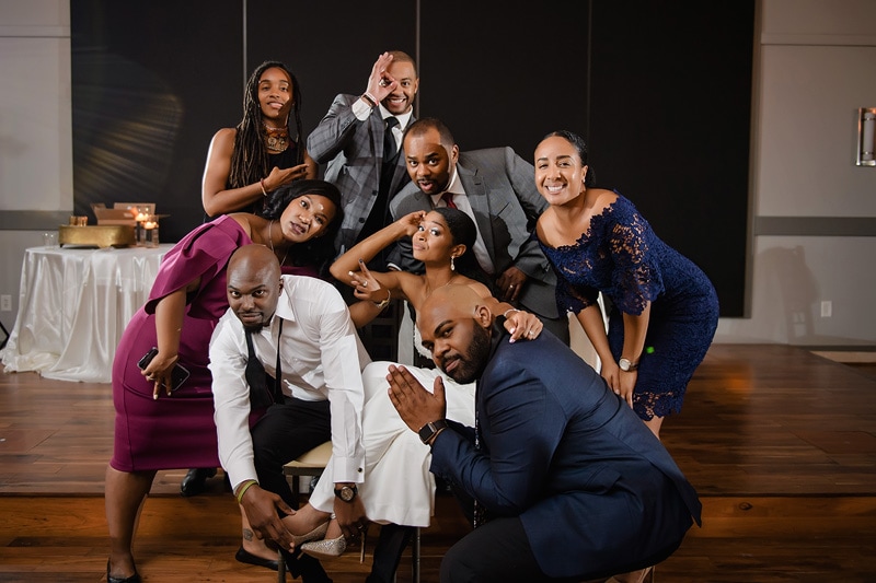 A group of nine elegantly dressed individuals pose cheerfully for a photo at a Paramount Event Venue wedding, showcasing their playful camaraderie and diverse personalities.