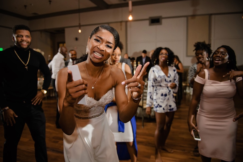 A joyful woman gives thumbs up while dancing at a Paramount Event Venue wedding, where guests are enjoying the moment, dancing and laughing in the background.