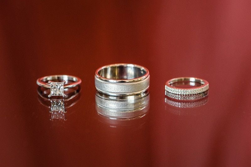 A trio of elegant wedding rings displayed on a glossy red surface at the Paramount Event Venue, featuring a solitaire diamond engagement ring and two bands with intricate designs, symbolizing love and partnership.
