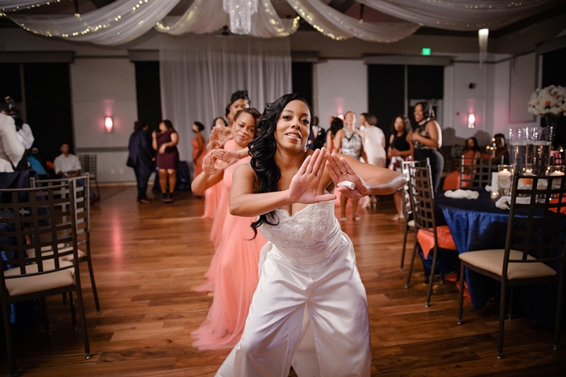 A joyful bride in a white dress dances enthusiastically with a bridesmaid in a coral gown at a lively wedding reception at Paramount Event Venue, surrounded by guests and festive decorations.