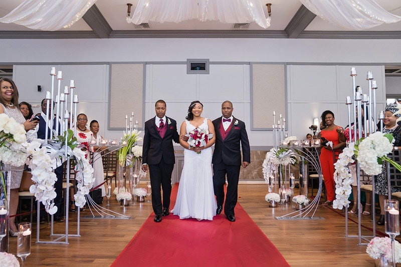 A joyful bride walks down the aisle, flanked by two smiling men, amid a beautifully decorated Paramount Event Venue with white flowers, candles, and a red carpet, while guests look on with happiness.