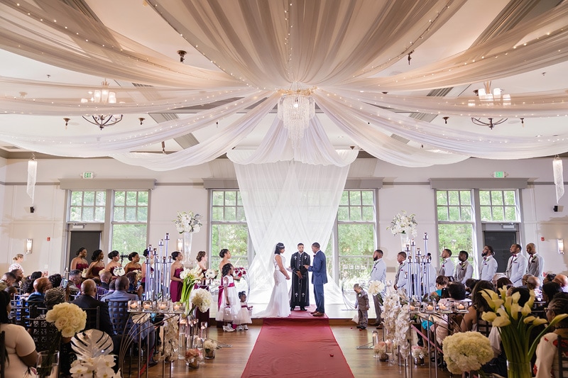A wedding ceremony in the Paramount Event Venue with elegant drapery, the bride and groom exchange vows at the altar, surrounded by bridesmaids, groomsmen, and seated guests witnessing the union.
