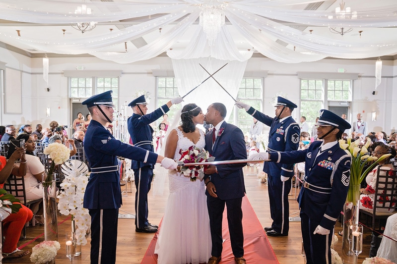 A military couple shares a kiss beneath an arch of sabers at their vibrant Paramount Event Venue wedding ceremony, surrounded by uniformed service members and guests in a festively decorated hall.