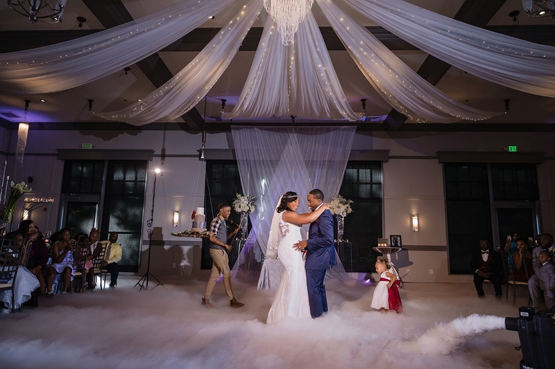 A newlywed couple shares their first dance at the Paramount Event Venue on a cloudlike dance floor, surrounded by guests and elegant decor, under a canopy of soft drapery and a chandelier.
