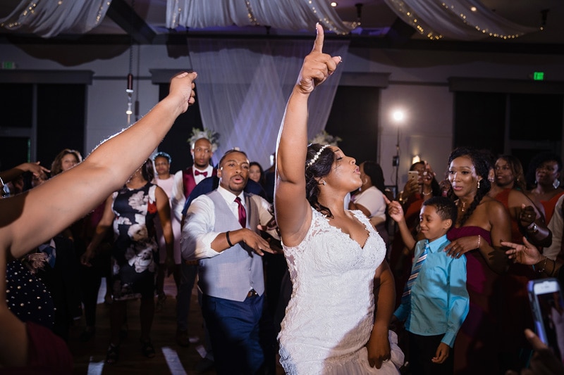 A joyful bride and groom dance at their Paramount Event Venue wedding reception, surrounded by cheering guests, with the bride raising her finger triumphantly as they celebrate their union.