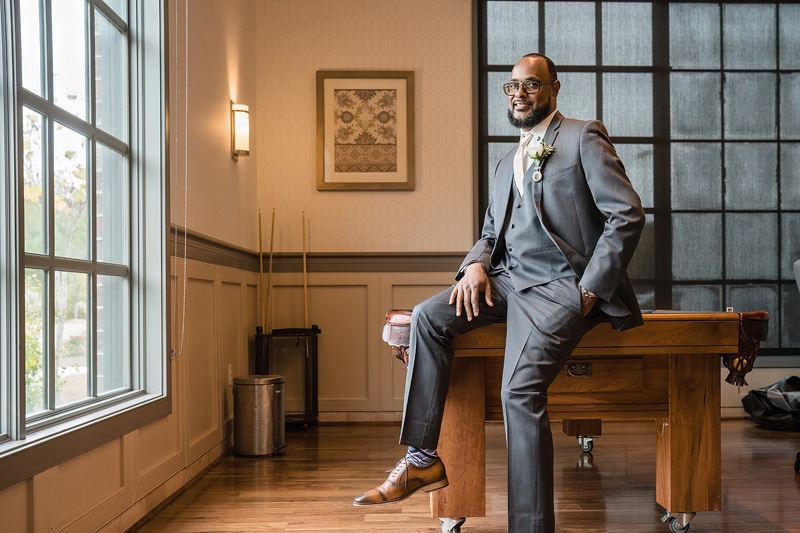 A distinguished man in a tailored suit with a boutonnière seated on a wooden desk, poised by a window in the Paramount Event Venue, projects confidence and elegance against an interior setting with subtle decor
