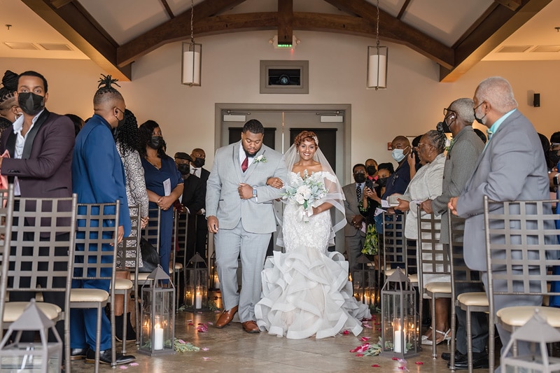 A joyful couple walks down the aisle together at the Paramount Event Venue, amidst the warm smiles of guests, the bride's gown trailing with elegance, and the aisle is adorned with rose petals and candles,