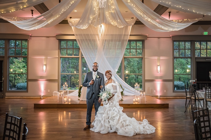 A beaming couple on their wedding day standing in the elegantly decorated Paramount Event Venue, with sheer draping, crystal chandeliers, and warm glowing candles, celebrating their union.