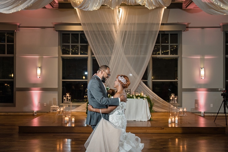 A newlywed couple shares a tender first dance beneath delicate drapery and twinkling lights, surrounded by the warm glow of candlelight in the Paramount Event Venue, exhibiting an elegant ballroom setting.