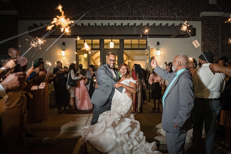 A joyful newlywed couple makes a grand exit under a sparkling archway of sparklers held by friends and family at the Paramount Event Venue, with expressions of happiness and celebration all around.