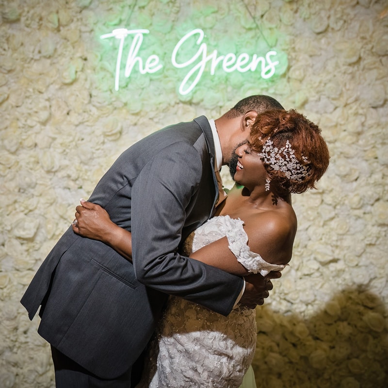 A joyful couple embraces on their wedding day at the Paramount Event Venue, illuminated by a neon sign bearing their surname, creating an intimate moment amid the festivity.