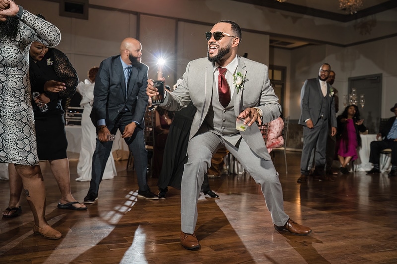 A joyous man in a dapper gray suit dances at a festive gathering at the Paramount Event Venue, drink in hand and a beaming smile, as onlookers join in the celebration.