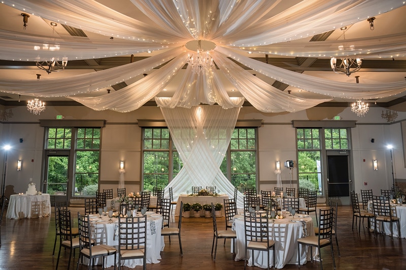 An elegantly decorated Paramount Event Venue banquet hall with draped ceiling fabric converging at a central chandelier, round tables set for guests, and a designated area with a greenery-adorned backdrop suggesting