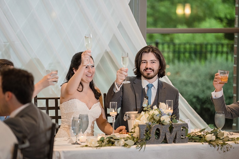 A radiant bride and groom joyfully raise their glasses for a toast at their wedding reception at the Paramount Event Venue, surrounded by guests and adorned with elegant decor and a sign that reads "love.