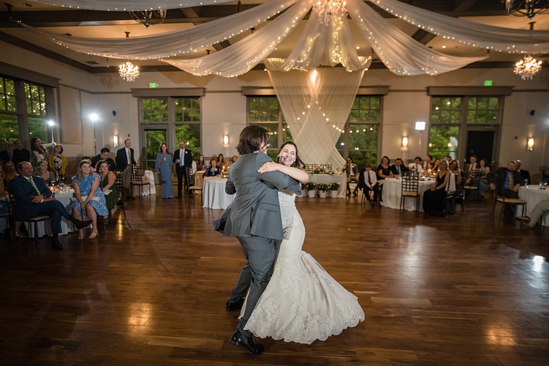 A joyful bride and groom share their first dance at the Paramount Event Venue, beneath elegant draped linens and twinkling lights, as wedding guests look on with smiles in a spacious, well-appointed venue.
