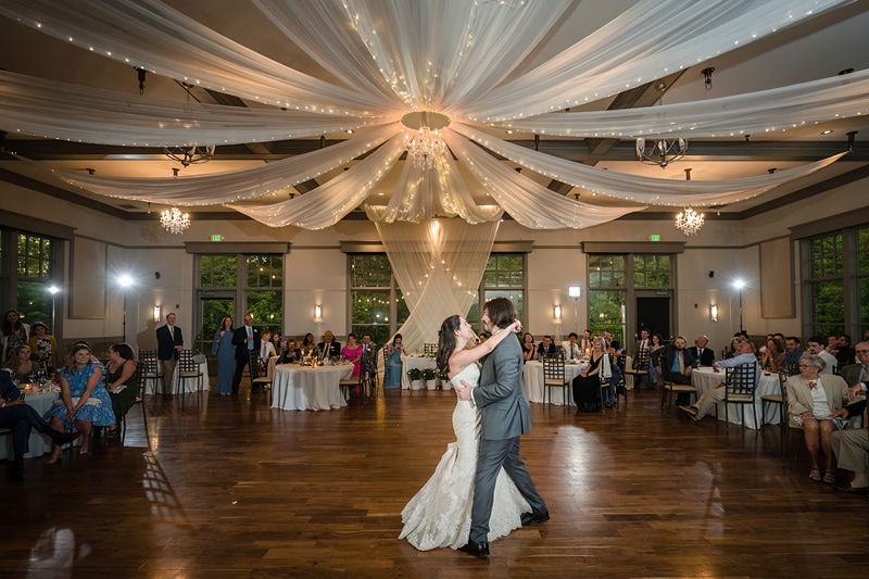 A bride and groom share their first dance at the Paramount Event Venue, an elegant hall adorned with draped linins and a chandelier, surrounded by seated guests watching on in celebration.
