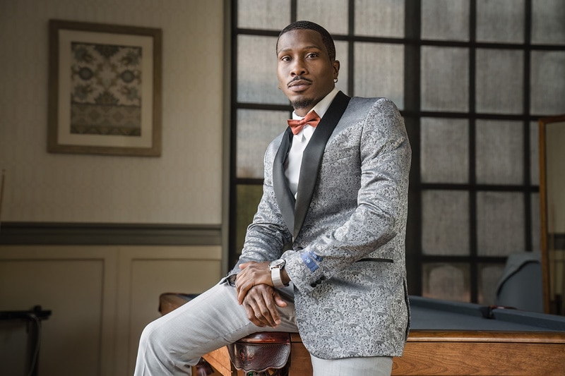 A confident man in a stylish grey patterned blazer and bow tie sits atop a wooden desk in a well-appointed room at the Paramount Event Venue, a picture of elegance and composure, ready for