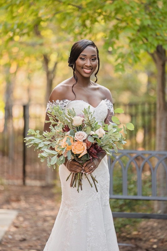 A radiant bride in an off-the-shoulder lace gown smiles gently, holding an elegant bouquet with peach roses and lush greenery, set against a tranquil, tree-lined path at the Paramount Event Venue