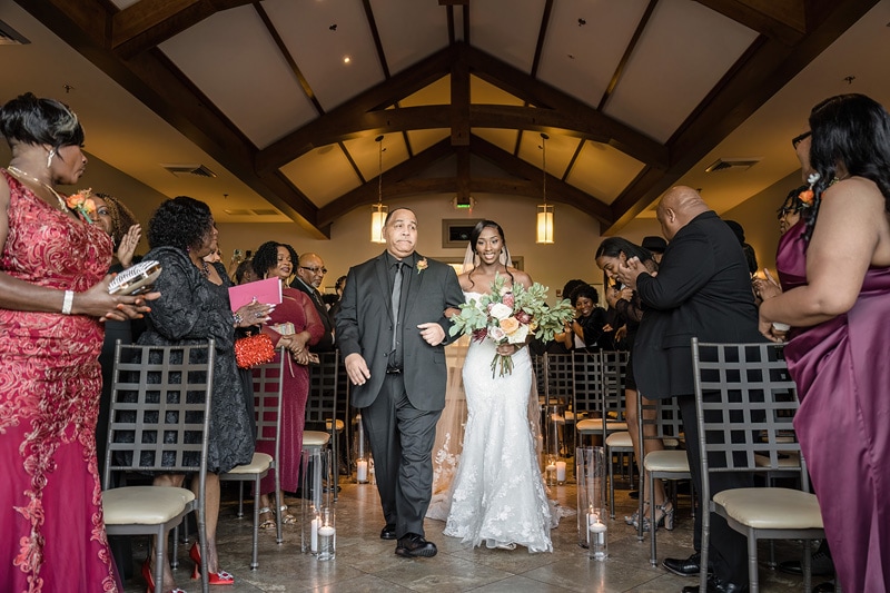 A radiant bride walks down the aisle at the Paramount Event Venue on the arm of a proud companion amidst an aisle of admiring guests, their attention captivated by her elegance, as soft lighting enhances the
