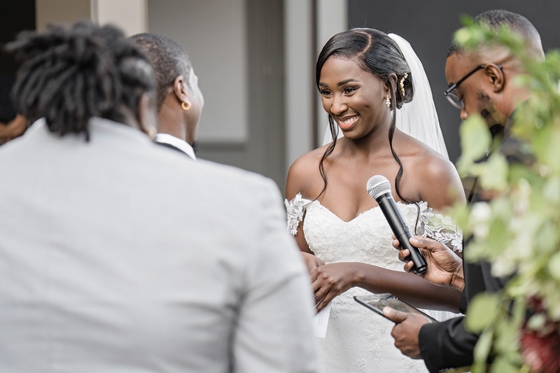 A radiant bride in a lace wedding dress shares heartfelt vows, displaying a joyful smile as she holds hands with her partner during a charming outdoor ceremony at the Paramount Event Venue, surrounded by attentive friends.