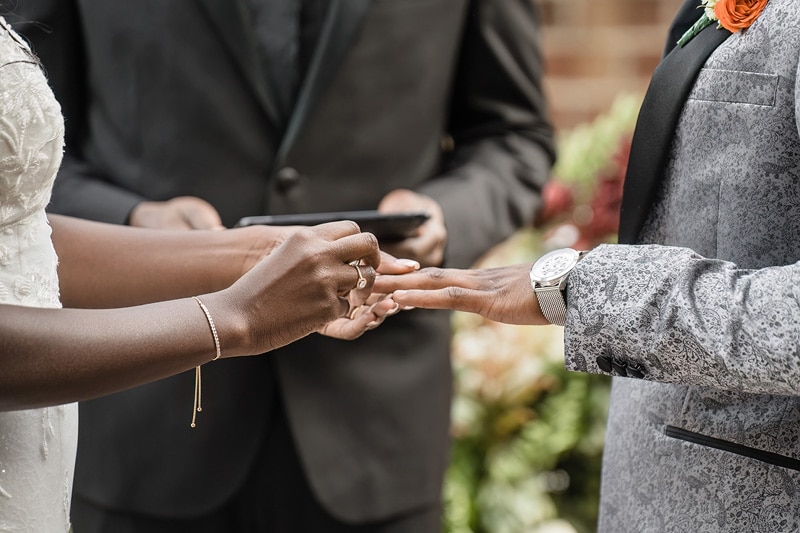 A bride and groom exchange wedding rings, a symbol of eternal love and commitment, during a ceremony at the Paramount Event Venue with the officiant in the background.