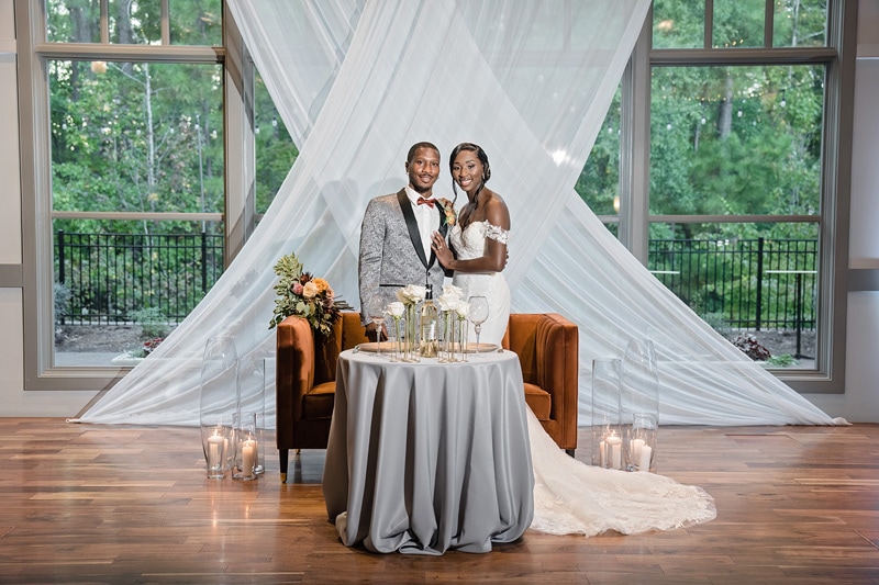 A newlywed couple poses elegantly at their Paramount Event Venue wedding reception, standing behind a round table adorned with candles and floral arrangements, with a sheer backdrop and lush greenery visible through large windows.