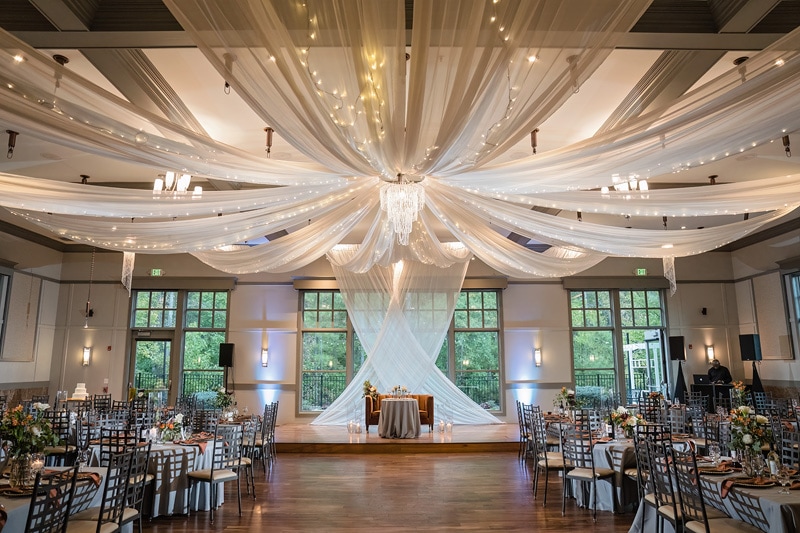 An elegantly decorated Paramount Event Venue set for a formal event, with tables adorned with centerpieces, a draped ceiling with warm lighting, and a designated area that could serve as a stage or focal point