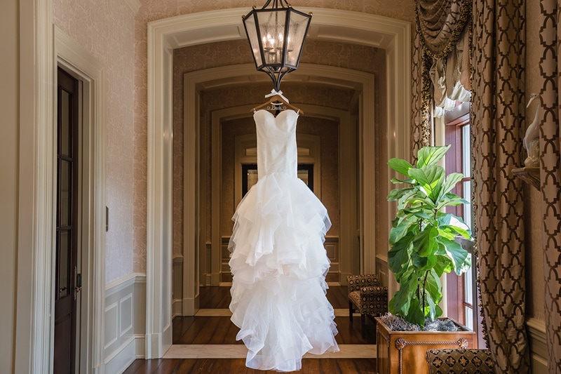 An elegant white wedding dress hangs in a luxurious hallway with wood flooring, draped curtains, and classic decor at Prestonwood Country Club, awaiting its moment in the spotlight.