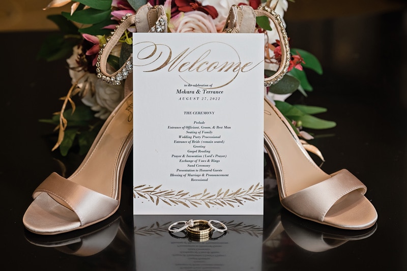 A Prestonwood Country Club wedding welcome card with details stands amidst a bouquet and satin bridal shoes, flanked by wedding rings, symbolizing the elegance and romance of marriage.